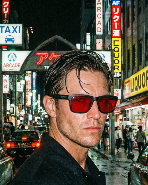 Man wearing red sunglasses in a neon-lit city street at night.